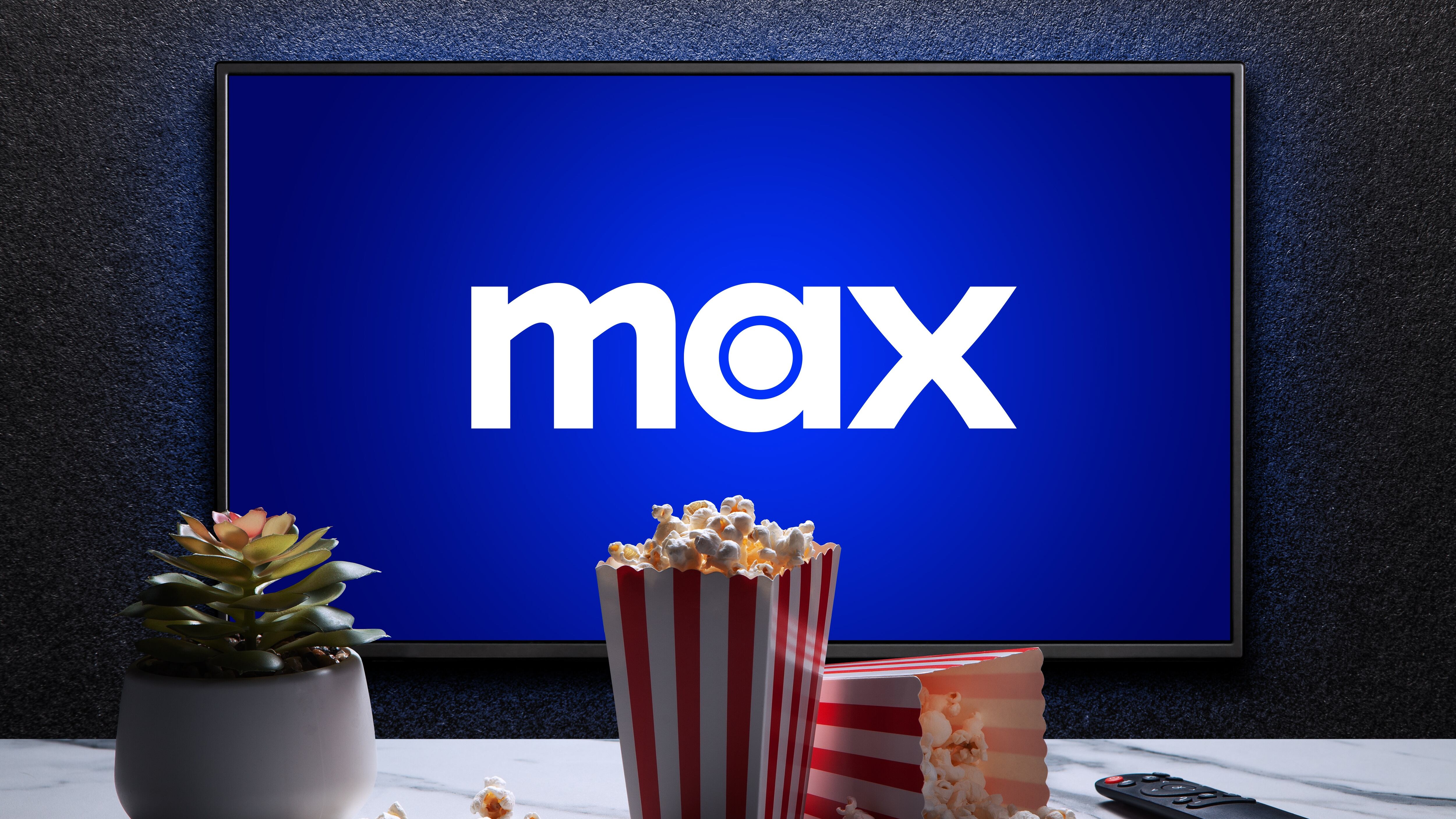 Max or HBO Max logo on TV with popcorn boxes, remote control and home plant on the table.